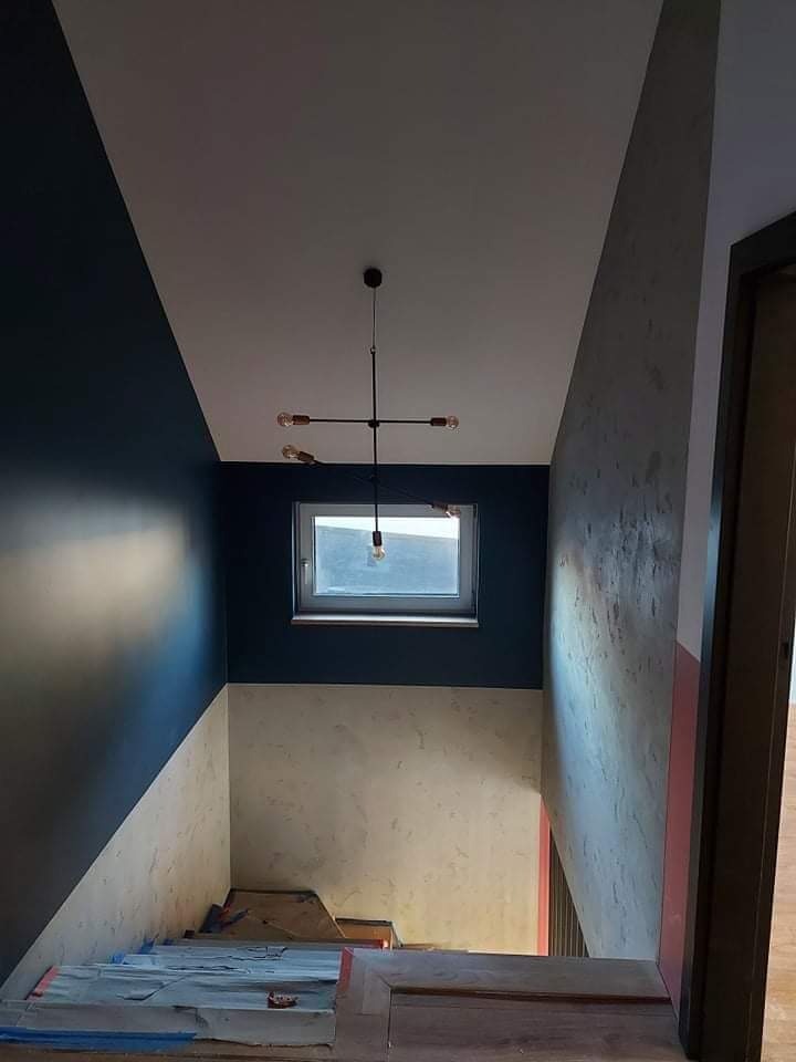 Klatka schodowa w trakcie remontu: ściany częściowo pomalowane na granatowo i pokryte dekoracyjnym tynkiem, zabezpieczone schody, nowoczesna lampa wisząca.