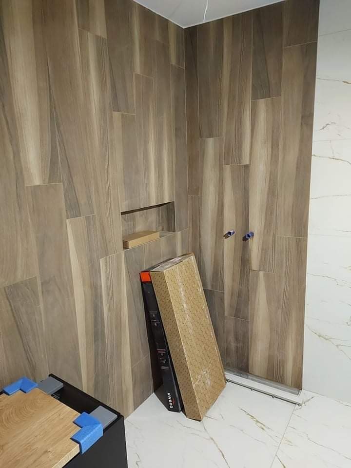 Łazienka w trakcie wykańczania: ściany wyłożone drewnopodobnymi płytkami w pionowym układzie, wnęka w ścianie, częściowo widoczne opakowania płytek i narzędzia na podłodze z marmurowym wzorem.