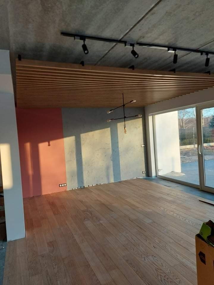 Wnętrze w trakcie wykańczania: drewniana podłoga, sufit z drewnianych listew, oświetlenie szynowe, ściana w odcieniach różu i szarości, duże okno przesuwne.