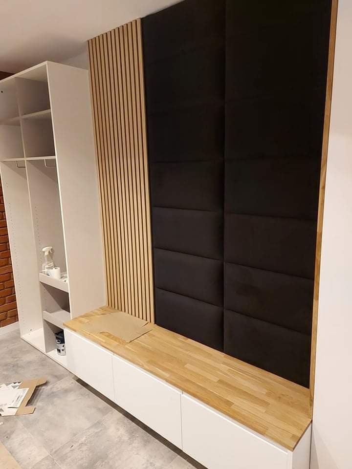 Wnętrze w trakcie montażu: biała szafa z półkami, drewniany blat, pionowe drewniane lamele i czarne panele tapicerowane na ścianie.