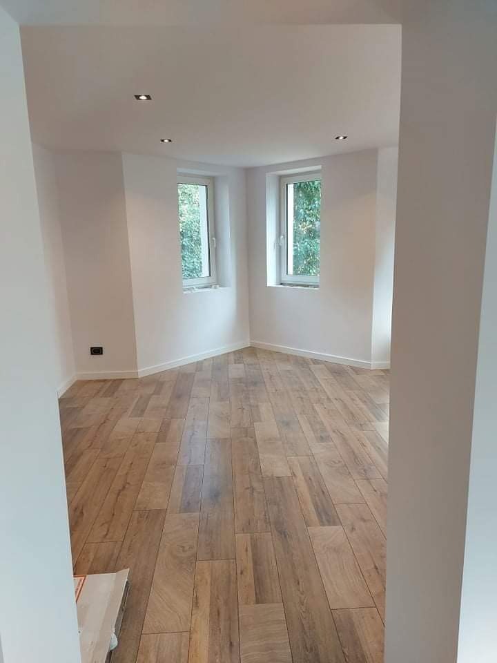 Jasne wnętrze po remoncie z drewnopodobną podłogą, białymi ścianami i dwoma oknami w narożniku, widoczne punktowe oświetlenie sufitowe.