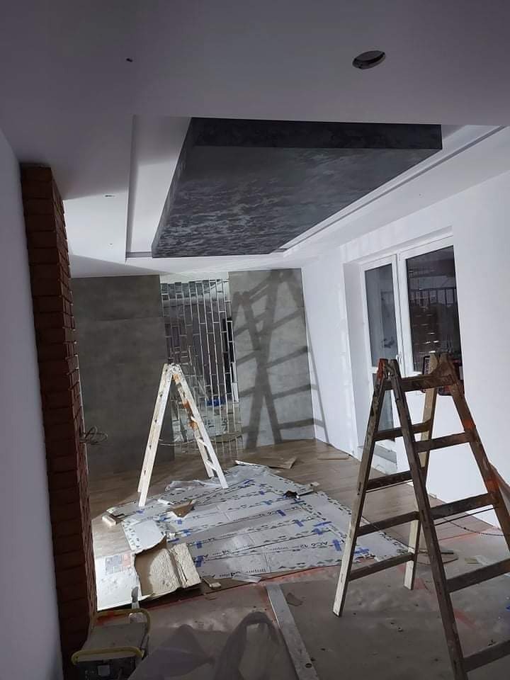 Wnętrze w trakcie prac wykończeniowych: widoczne drabiny, zabezpieczona podłoga, ściany przygotowane pod malowanie, dekoracyjny sufit podwieszany z ciemną, strukturalną powierzchnią.