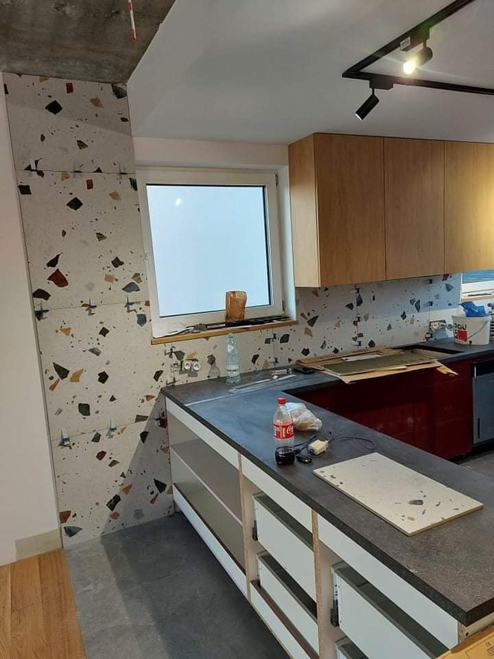 Montaż płytek imitujących lastryko na ścianie w kuchni, widoczny surowy betonowy sufit i niedokończona zabudowa meblowa z szarym blatem.