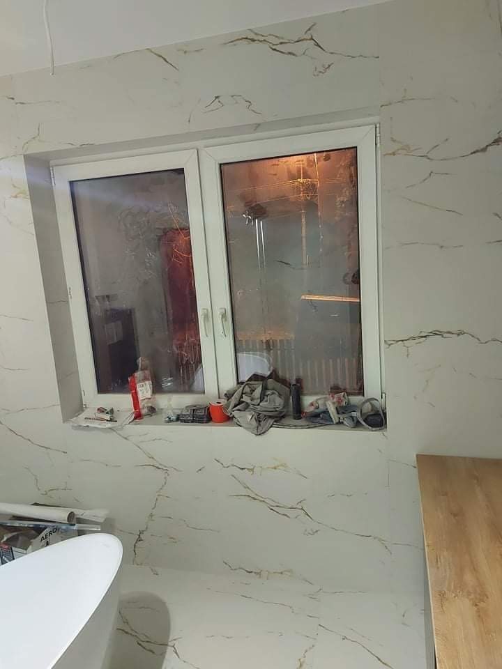 Łazienka w trakcie remontu z białymi płytkami imitującymi marmur, widok na okno z narzędziami na parapecie i częściowo widoczną wannę.