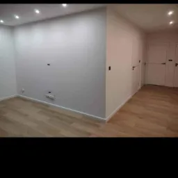 Jasne wnętrze po remoncie: świeżo pomalowane ściany w odcieniu bieli, panele podłogowe w kolorze jasnego drewna, trzy białe drzwi z czarnymi klamkami, oświetlenie punktowe w suficie.