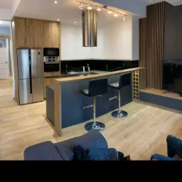 Nowoczesna kuchnia z wyspą i dwoma hokerami, jasne drewno i białe szafki, stalowy okap i podłużne oświetlenie sufitowe, widok z salonu.