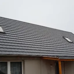 Dom jednorodzinny z ciemnoszarą dachówką, dwoma oknami dachowymi i drewnianą wiatą przy wejściu, widok z dołu w pochmurny dzień.