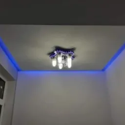 Pomieszczenie z oświetleniem LED wzdłuż krawędzi sufitu i ozdobnym żyrandolem z kryształkami, widok z dołu na gładkie, białe ściany i okno.