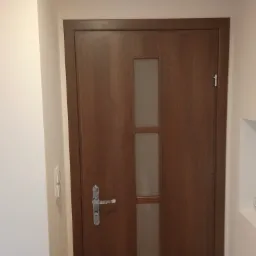 Zamknięte, drewniane drzwi wewnętrzne z przeszkleniem, zamontowane w jasnym wnętrzu w Szlichtyngowej. Widoczna srebrna klamka i włącznik światła na ścianie.