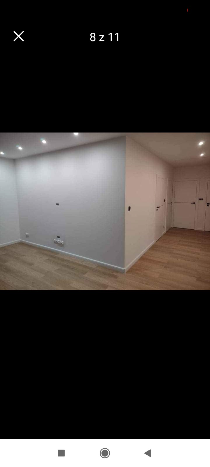 Jasne wnętrze po remoncie: świeżo pomalowane ściany w odcieniu bieli, panele podłogowe w kolorze jasnego drewna, trzy białe drzwi z czarnymi klamkami, oświetlenie punktowe w suficie.