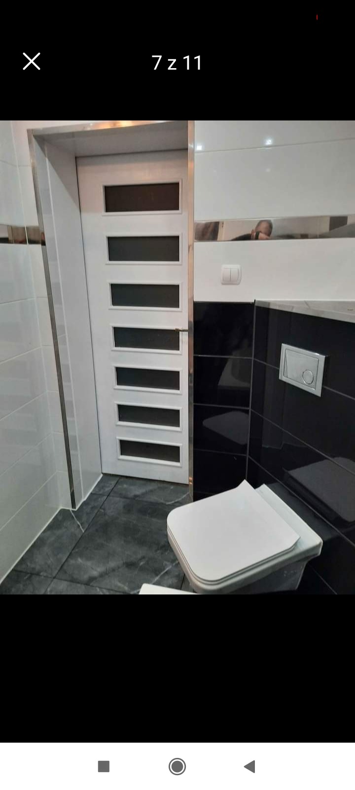 Wykończona łazienka z białą, przeszkloną, wewnętrzną stolarką drzwiową, czarno-białymi płytkami na ścianach i szarą podłogą z płytek imitujących marmur.