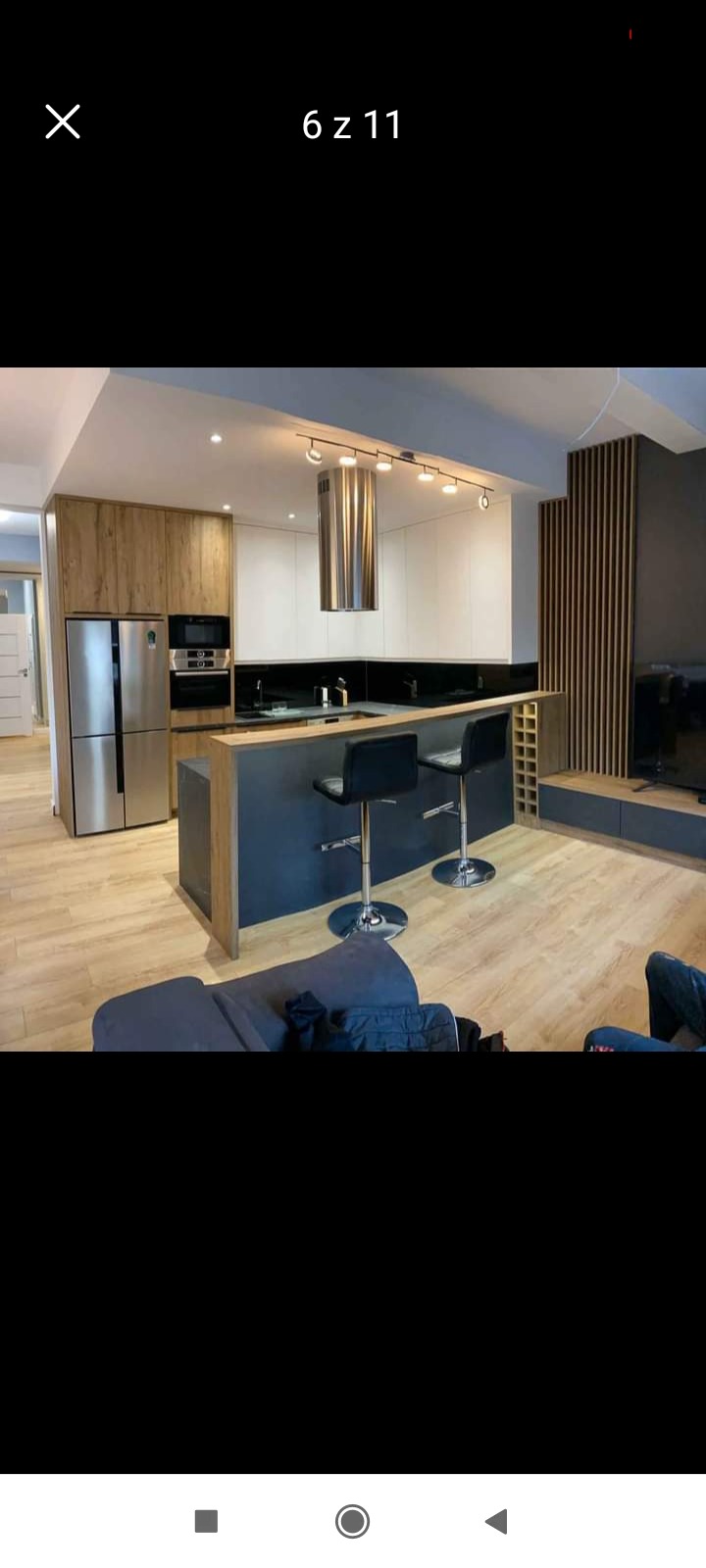 Nowoczesna kuchnia z wyspą i dwoma hokerami, jasne drewno i białe szafki, stalowy okap i podłużne oświetlenie sufitowe, widok z salonu.