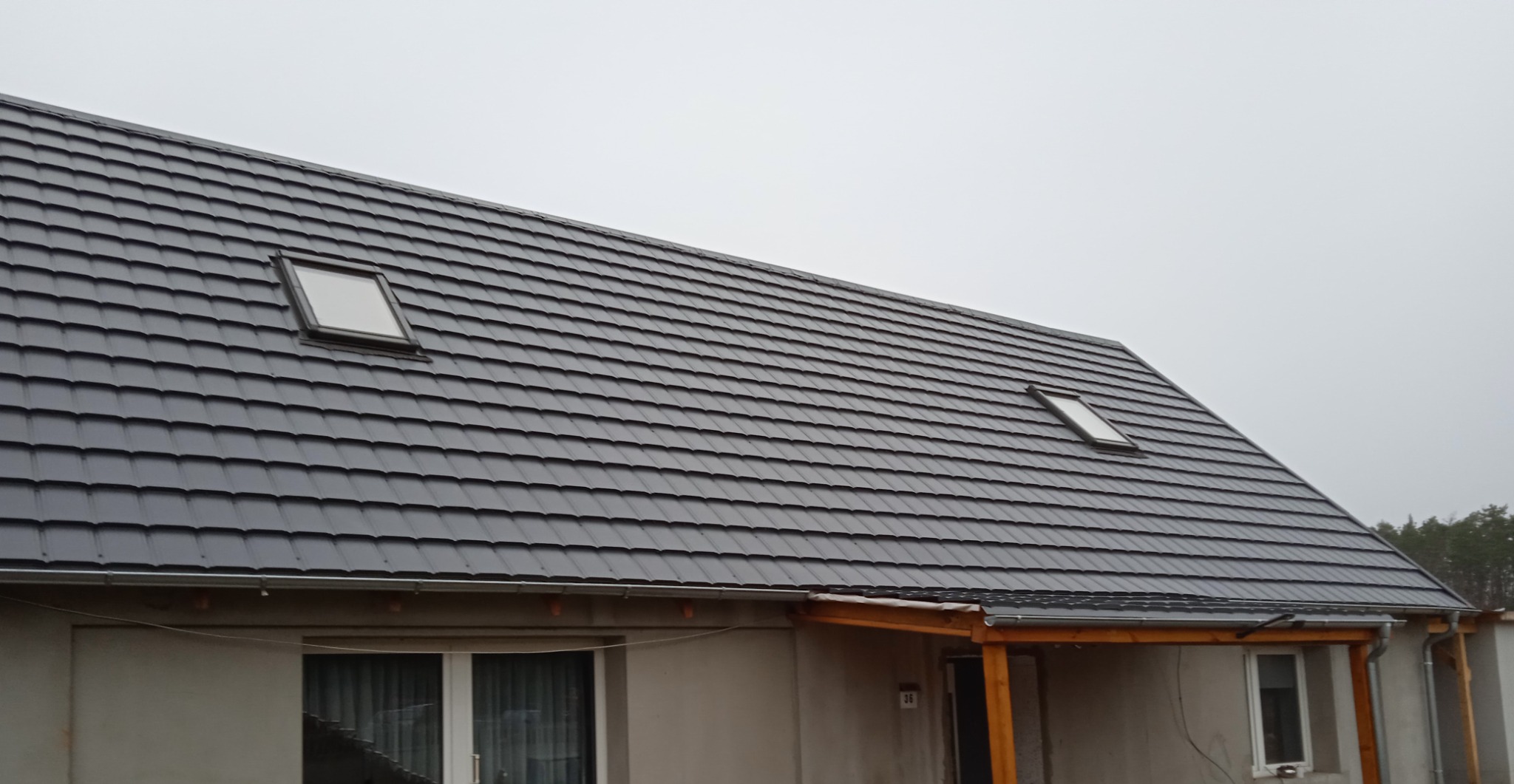 Dom jednorodzinny z ciemnoszarą dachówką, dwoma oknami dachowymi i drewnianą wiatą przy wejściu, widok z dołu w pochmurny dzień.