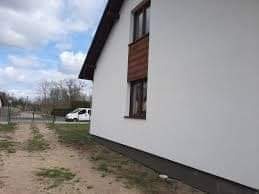 Świeżo otynkowana, biała ściana zewnętrzna budynku jednorodzinnego z brązowymi ramami okiennymi, widoczna z perspektywy gruntu, z fragmentem podjazdu i samochodem w tle.