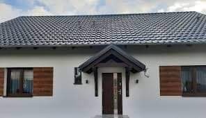 Biała elewacja domu jednorodzinnego z ciemnoszarą dachówką, brązowymi okiennicami i zadaszeniem nad wejściem. Widoczne rynny i oświetlenie zewnętrzne.