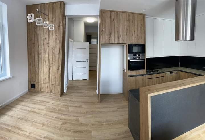 Nowoczesna kuchnia z drewnianymi frontami szafek, czarnym blatem i zabudowanym sprzętem AGD, widok na przedpokój z białymi drzwiami.