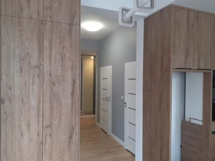 Nowoczesne wnętrze z zabudową meblową w odcieniach drewna, białe drzwi z poziomymi frezami, szare ściany i jasna podłoga, widok na korytarz z oświetleniem sufitowym.