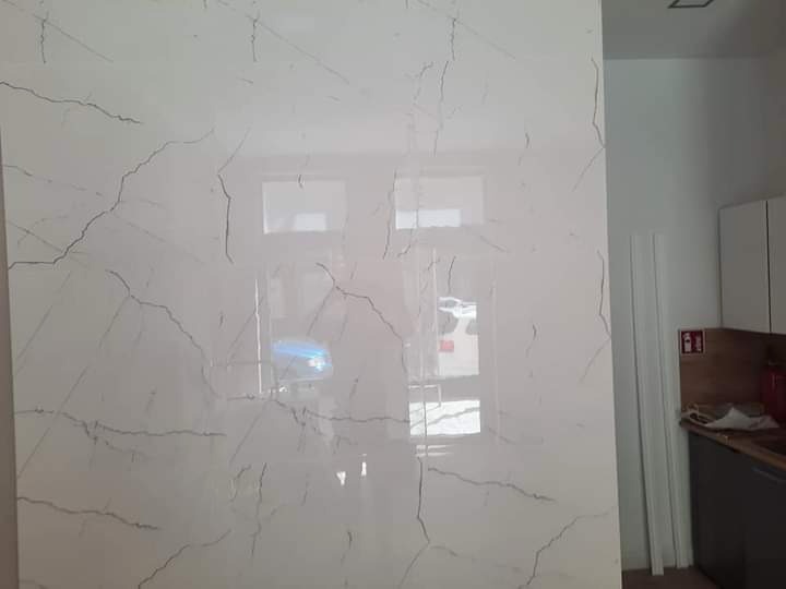 Błyszcząca ściana wykończona płytami imitującymi biały marmur z szarymi żyłami, odbijająca światło z okna, widoczny fragment blatu kuchennego i szafek.