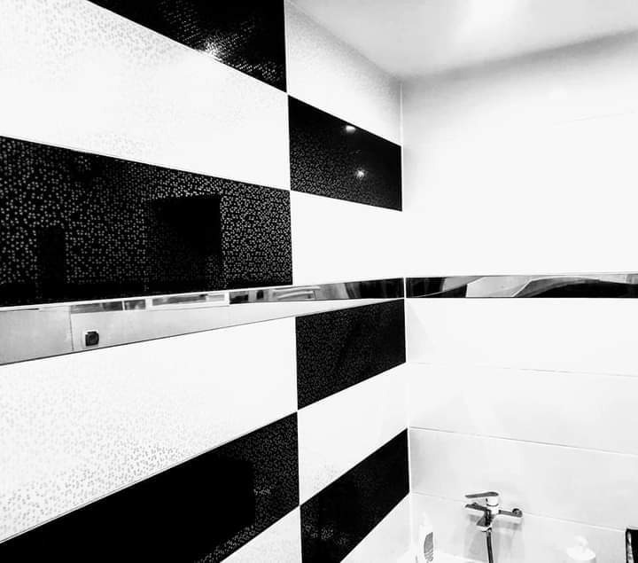 Czarno-biała mozaika w szachownicę na ścianie łazienki, wykończona metalową listwą, z widoczną baterią umywalkową i fragmentem blatu.