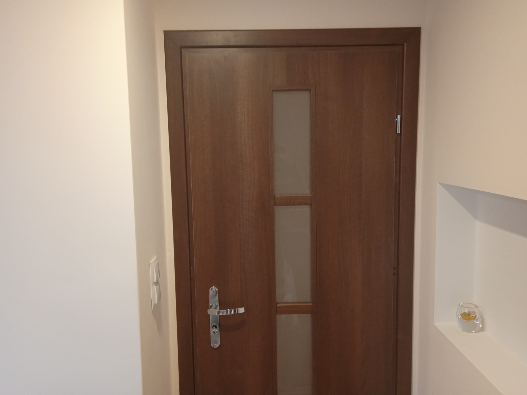 Zamknięte, drewniane drzwi wewnętrzne z przeszkleniem, zamontowane w jasnym wnętrzu w Szlichtyngowej. Widoczna srebrna klamka i włącznik światła na ścianie.