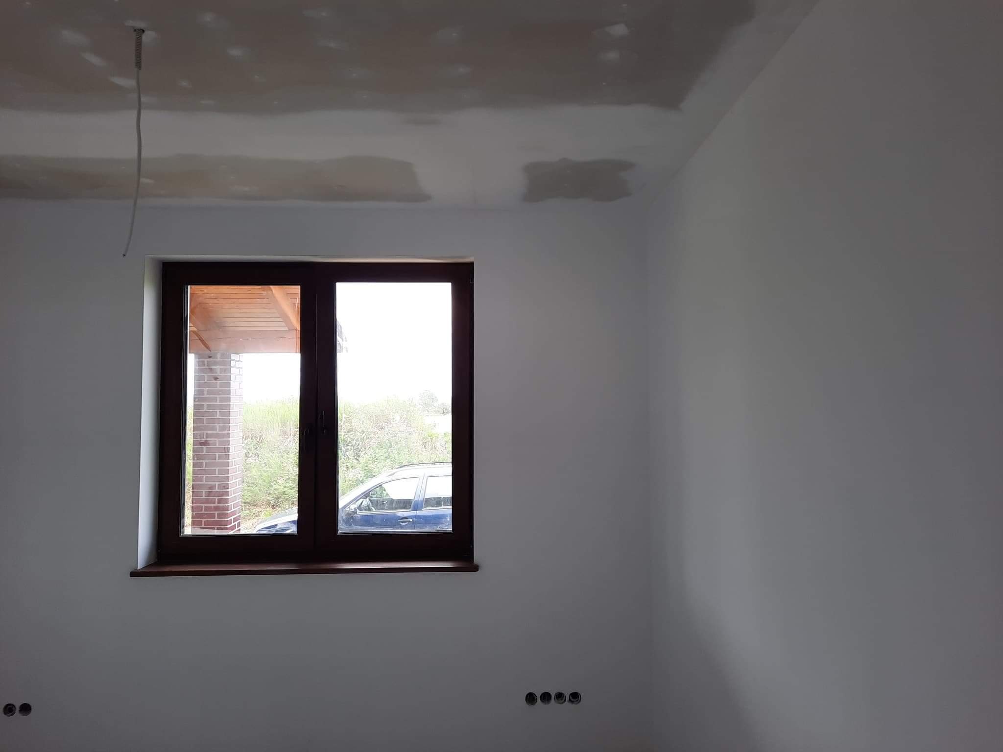 Wnętrze pokoju w trakcie prac wykończeniowych, widoczne ślady szpachlowania na suficie, białe ściany, brązowe okno z widokiem na zewnątrz.