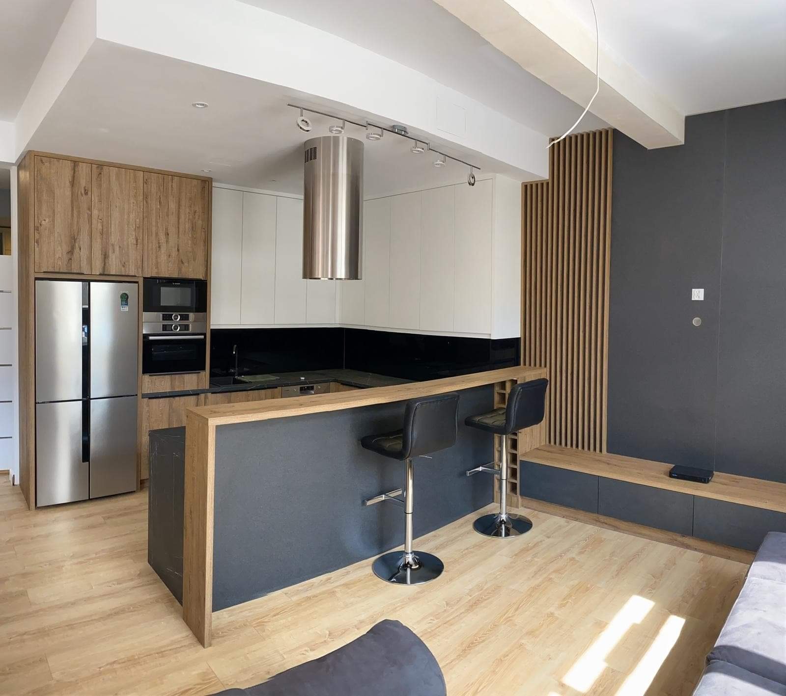 Nowoczesna kuchnia z wyspą, drewnianymi i białymi szafkami, chromowanym okapem cylindrycznym i barkiem z dwoma hokerami na tle szarej ściany z drewnianymi lamelami.