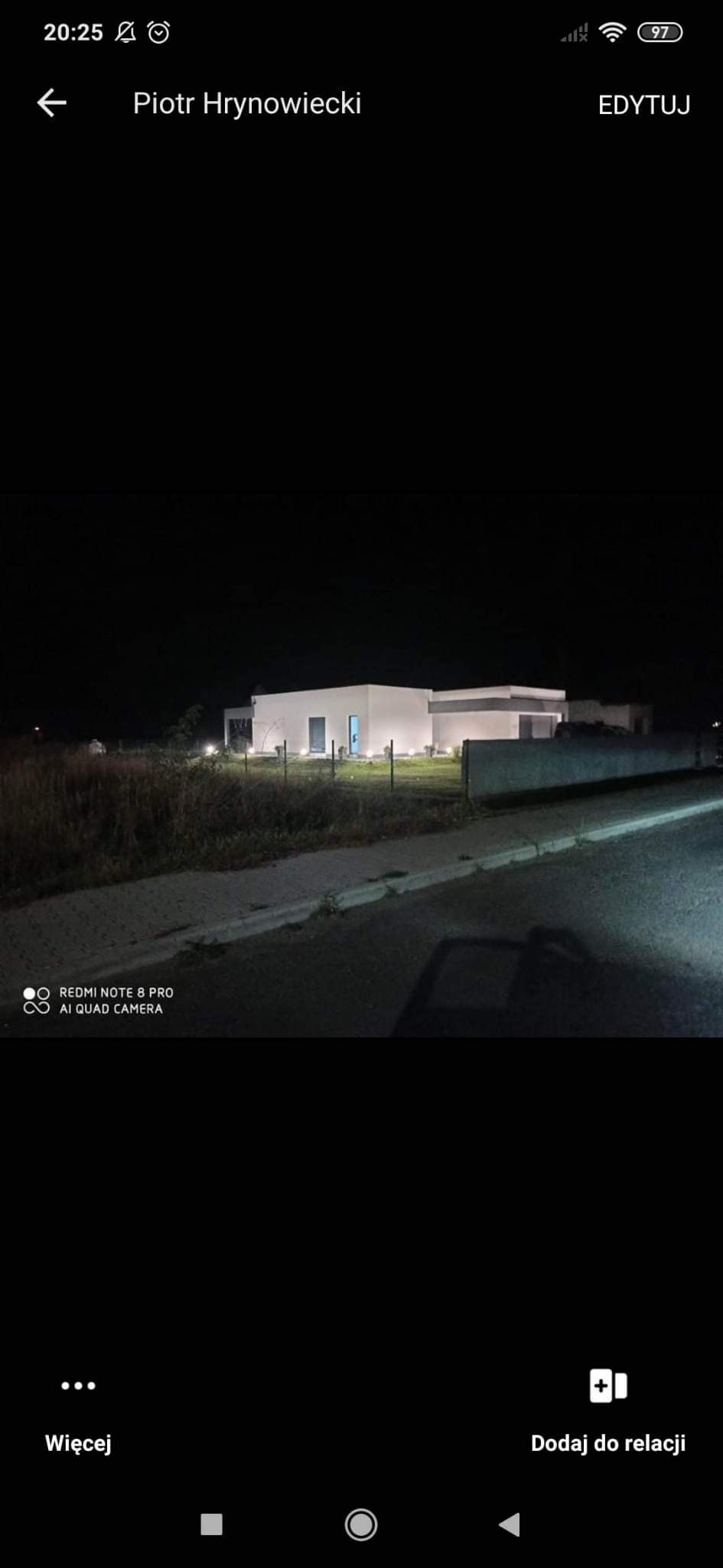 Nocne ujęcie nowo wybudowanego, parterowego domu z płaskim dachem, oświetlonego zewnętrznymi lampami, widoczne trawnik i ogrodzenie.