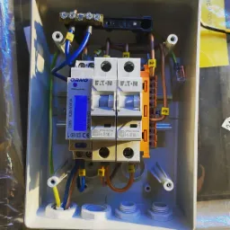 Rozdzielnica elektryczna w obudowie, widoczne bezpieczniki Eaton, licznik energii Orno z podświetlanym wyświetlaczem, listwa zaciskowa, przewody oznaczone numerami, uchwyty do mocowania na ścianie.