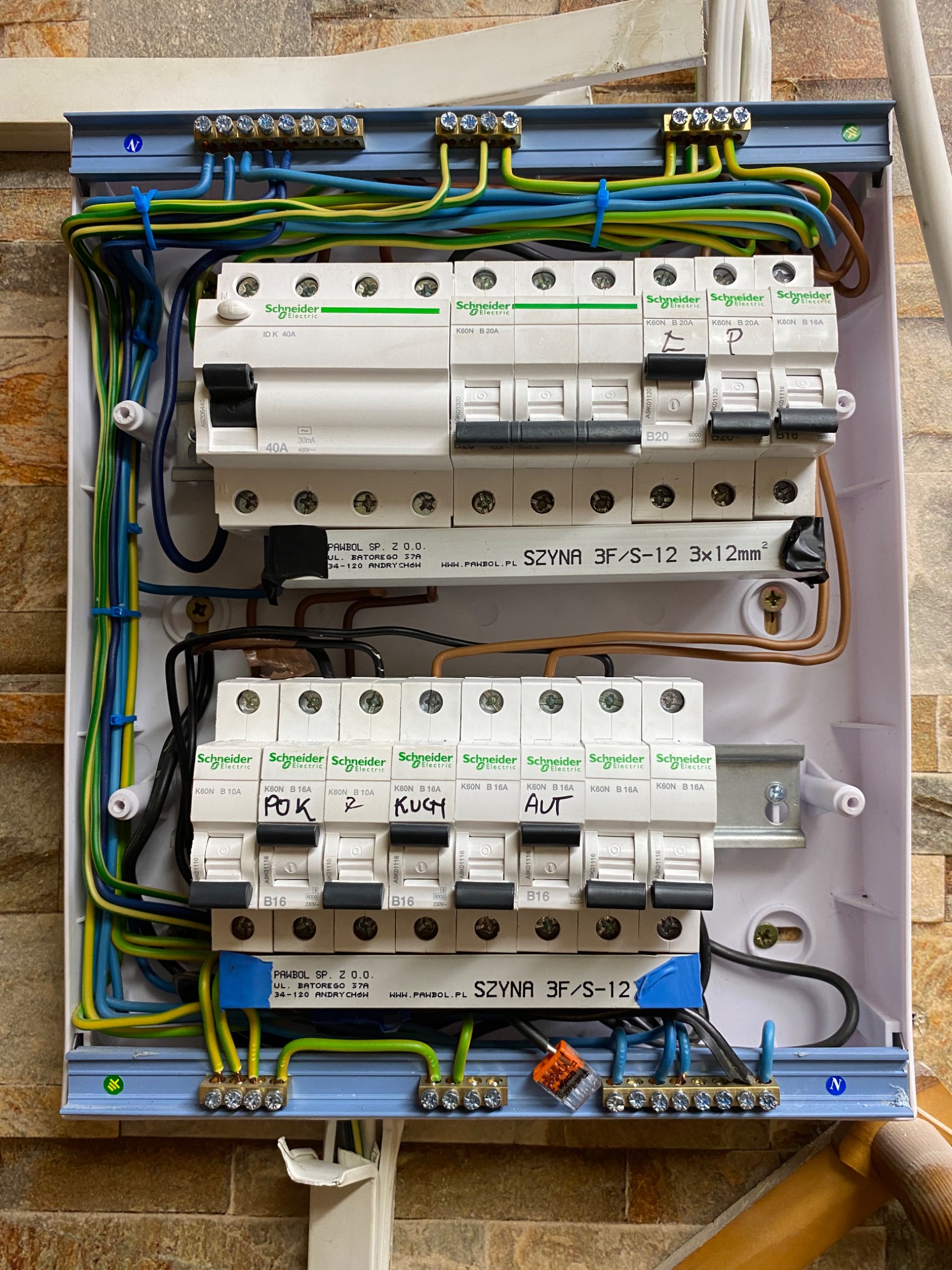 Otwarta skrzynka rozdzielcza z zamontowanymi bezpiecznikami Schneider Electric, okablowaniem w kolorach niebieskim, żółto-zielonym i brązowym, szynami PAWBOL, widoczne oznaczenia na bezpiecznikach...