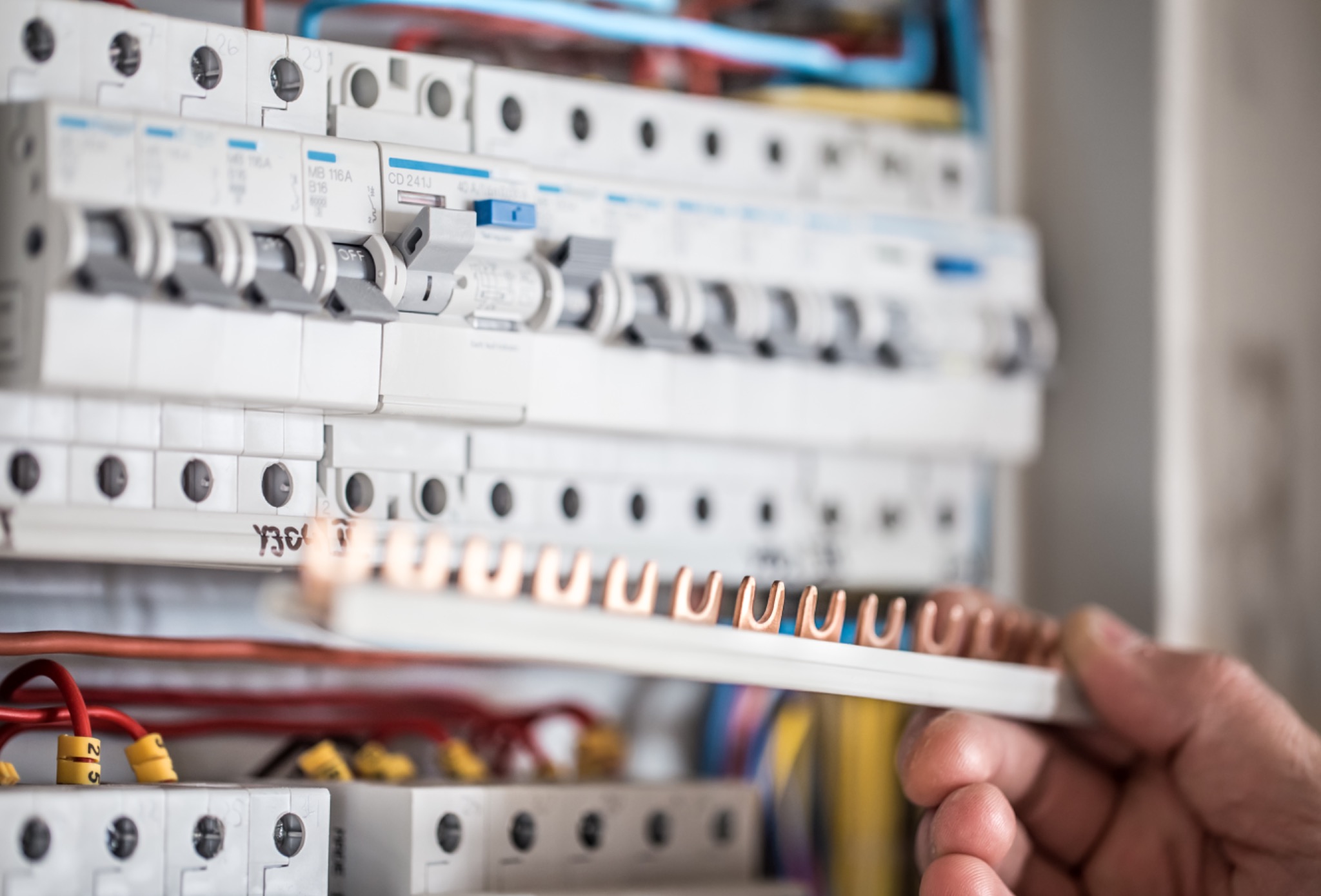 Otwarta skrzynka elektryczna z widocznymi bezpiecznikami i przewodami, ręka elektryka umieszcza listwę zaciskową z miedzianymi wsuwkami.
