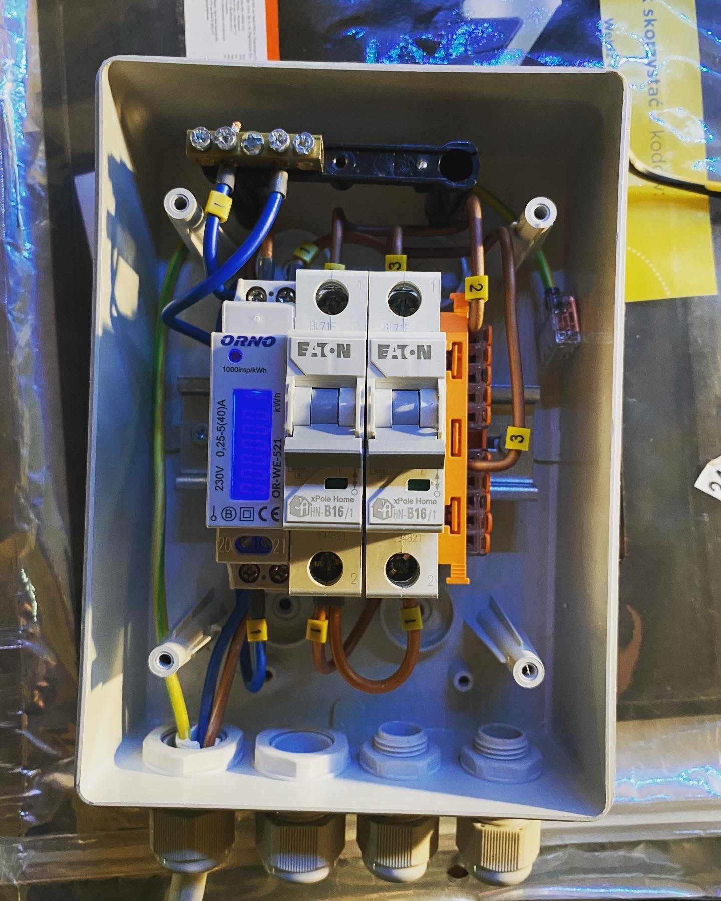 Rozdzielnica elektryczna w obudowie, widoczne bezpieczniki Eaton, licznik energii Orno z podświetlanym wyświetlaczem, listwa zaciskowa, przewody oznaczone numerami, uchwyty do mocowania na ścianie.
