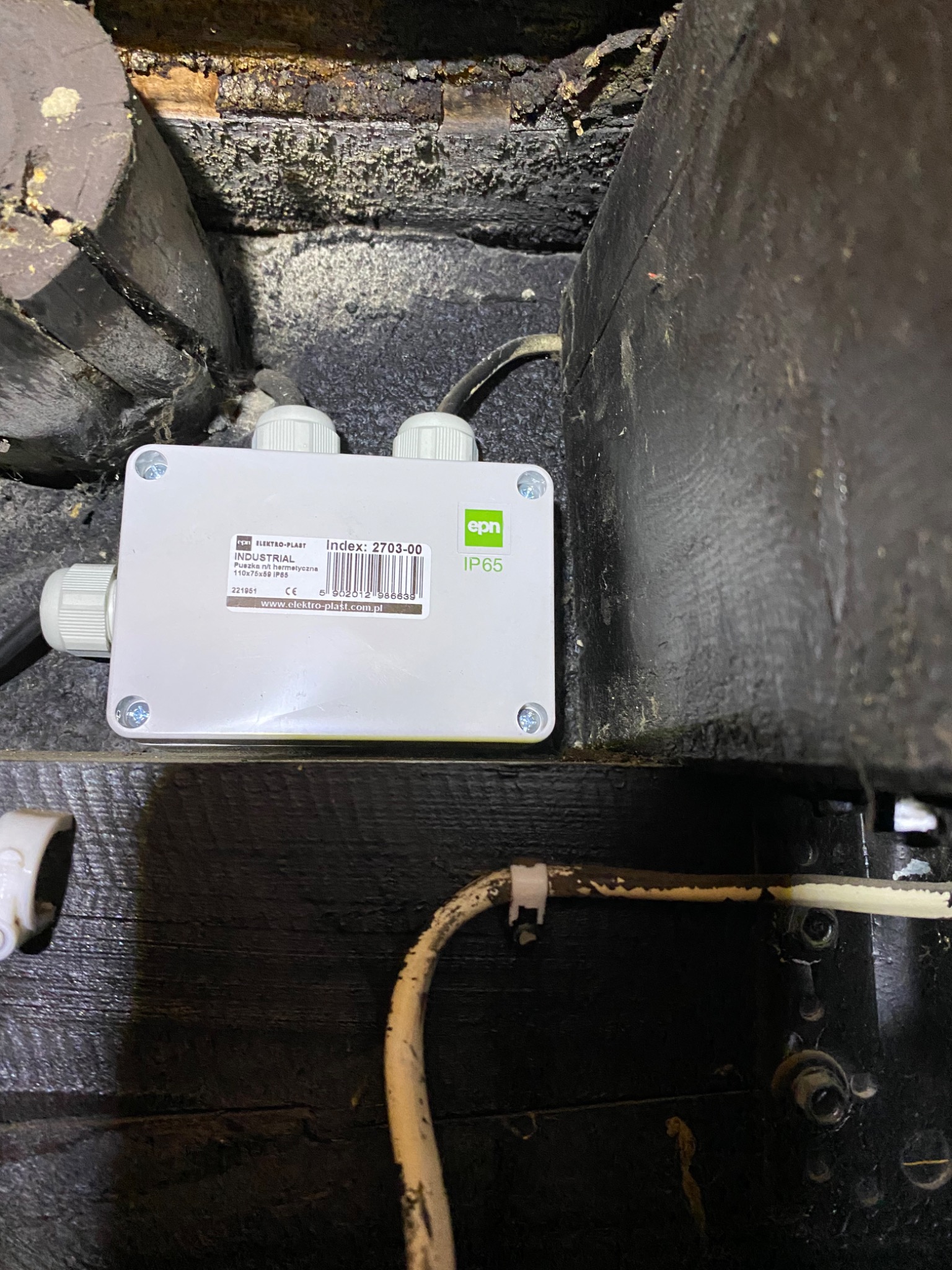 Biała puszka instalacyjna hermetyczna Elektro-Plast IP65, zamocowana na ciemnym, prawdopodobnie drewnianym podłożu z widocznymi kablami i elementami mocującymi.