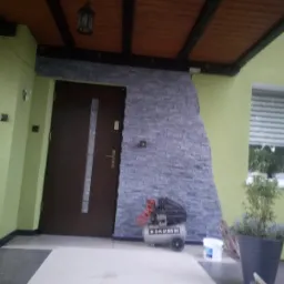 Wejście do domu z nową elewacją z kamienia dekoracyjnego, widoczny kompresor, donica z rośliną i drewniany sufit z lampą.