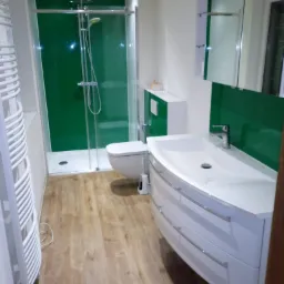 Nowoczesna łazienka z kabiną prysznicową z zielonym panelem, białą umywalką z szafką z szufladami, toaletą i grzejnikiem drabinkowym.