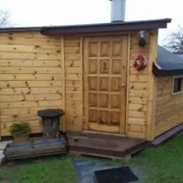 Drewniana konstrukcja z drzwiami, małym okienkiem i kominem, prawdopodobnie sauna ogrodowa, z dekoracyjnym wieńcem na drzwiach, stojąca na trawniku.