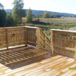 Świeżo wybudowany drewniany taras z balustradą i schodami, widok na wiejskie pole i las w oddali, jasne światło słoneczne tworzy wzory cieni na deskach podłogowych.