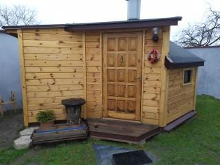 Drewniana konstrukcja z drzwiami, małym okienkiem i kominem, prawdopodobnie sauna ogrodowa, z dekoracyjnym wieńcem na drzwiach, stojąca na trawniku.