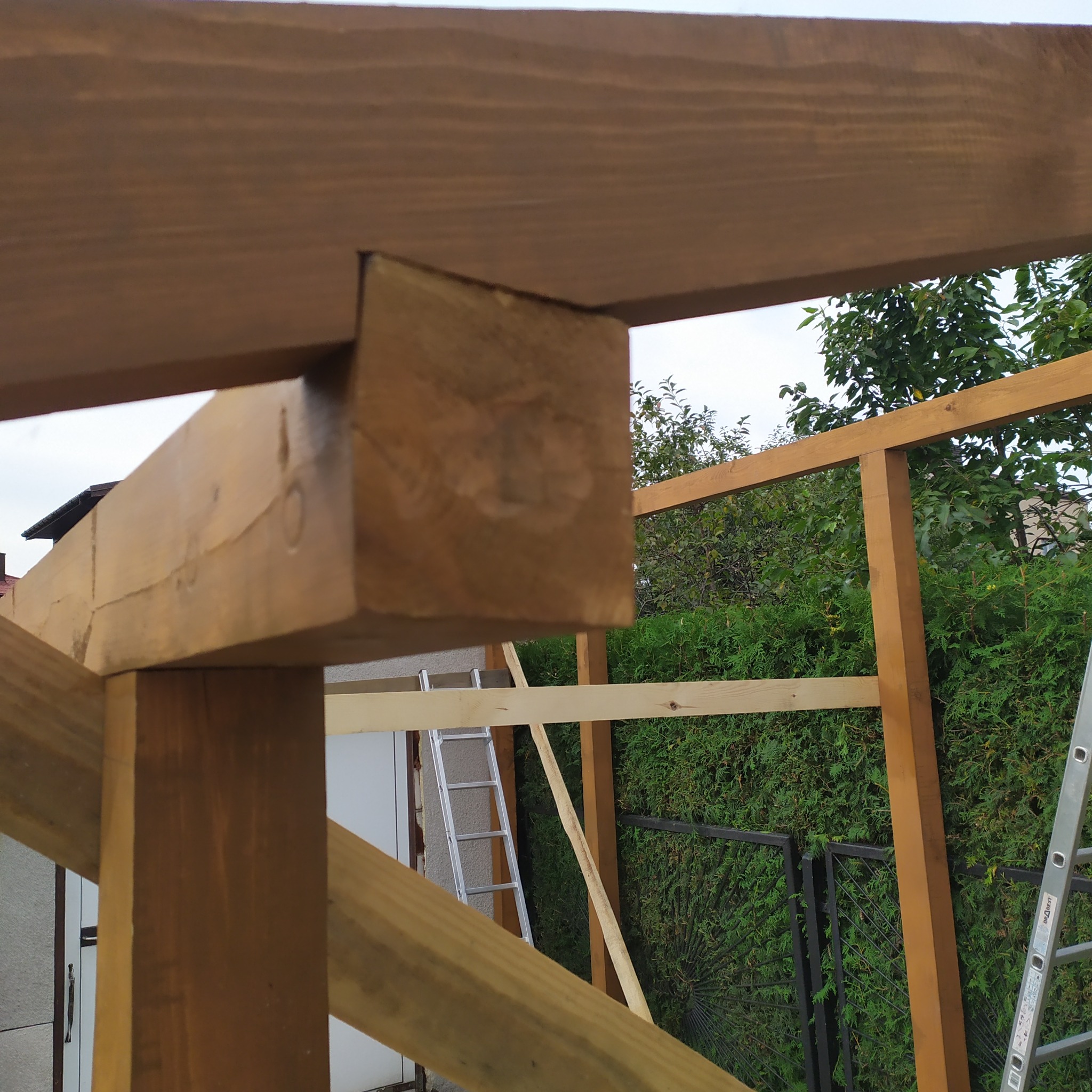 Fragment drewnianej konstrukcji w trakcie budowy, widoczne połączenia belek, drabina oparta o ścianę budynku w tle.