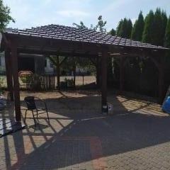 Drewniana konstrukcja zadaszona w Łomży z ciemnym dachem, rzucająca cień na brukowaną powierzchnię. Widoczne krzesło i puszka z farbą, sugerujące etap wykończeniowy.