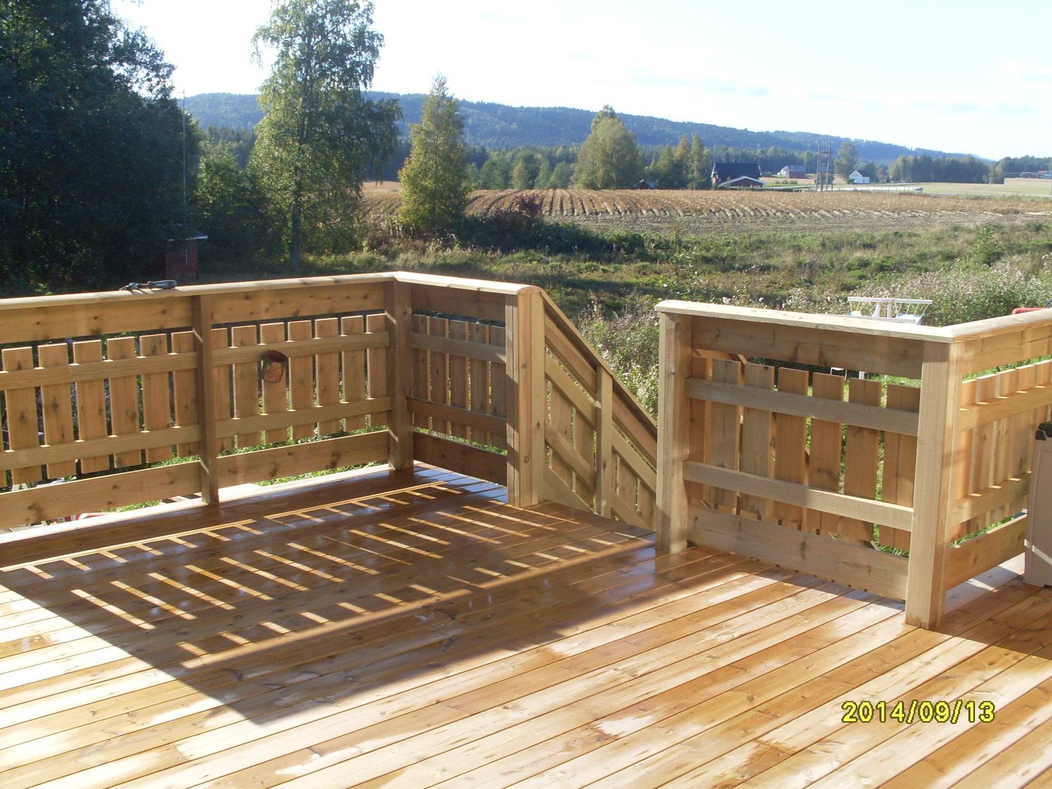 Świeżo wybudowany drewniany taras z balustradą i schodami, widok na wiejskie pole i las w oddali, jasne światło słoneczne tworzy wzory cieni na deskach podłogowych.