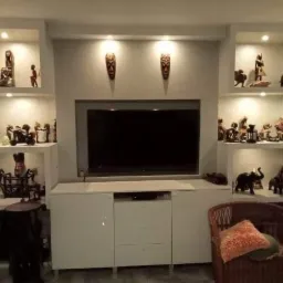 Biała zabudowa meblowa z wnękami oświetlonymi punktowo, wypełnionymi afrykańskimi rzeźbami i figurkami, zlokalizowana w Legionowie, z centralnie umieszczonym telewizorem i dekoracyjnymi maskami...