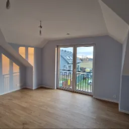 Jasne poddasze z drewnianą podłogą i świeżo pomalowanymi ścianami na delikatny odcień błękitu. Widoczne okno balkonowe z widokiem na sąsiedni dom i ogród. Na suficie widoczne niedokończone...