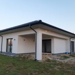 Nowoczesny dom jednorodzinny z białymi ścianami, czarnymi oknami i dachem, widok z zewnątrz na elewację, częściowo wykończony teren wokół budynku.