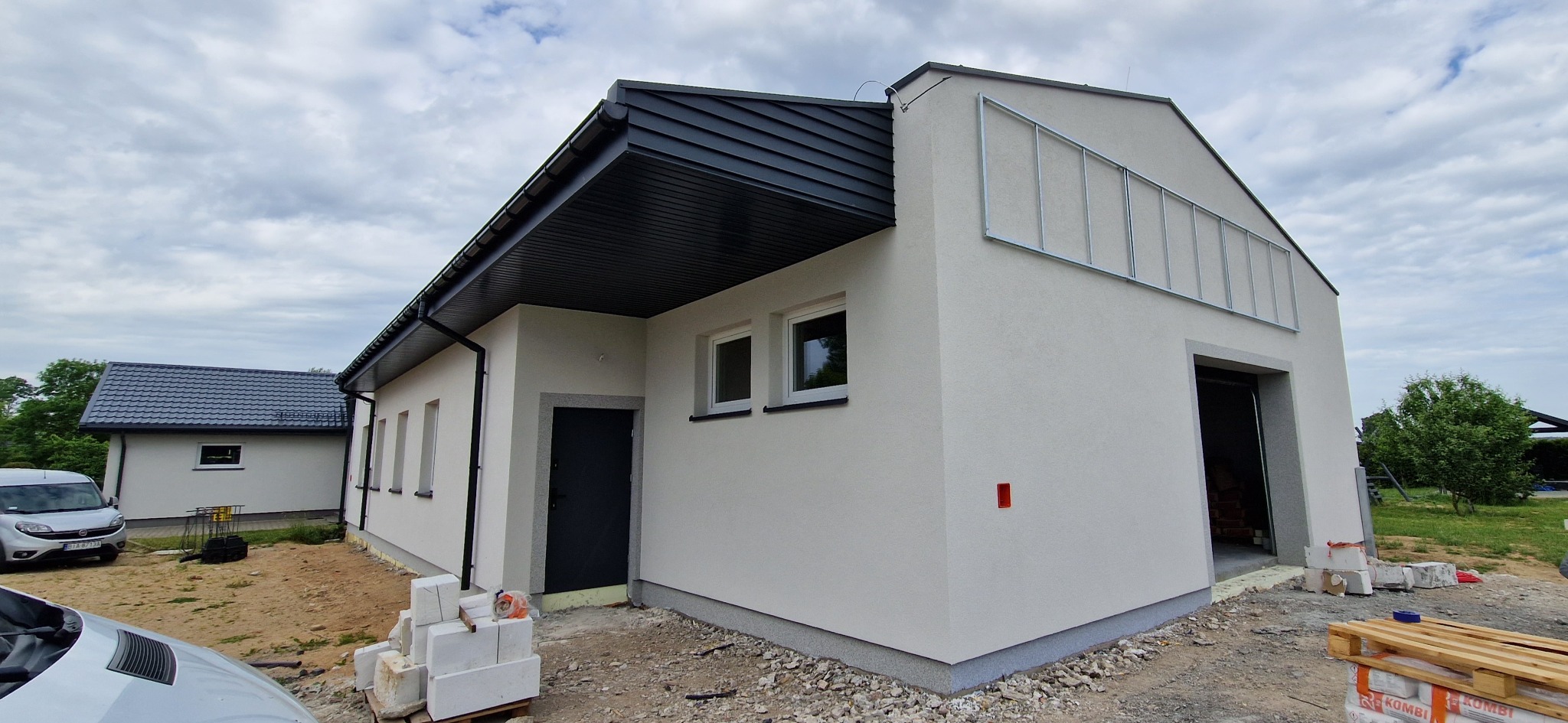 Nowo otynkowany budynek z ciemną blachodachówką i wykończonymi oknami, widoczne materiały budowlane i niezakończone prace wykończeniowe wokół budynku.