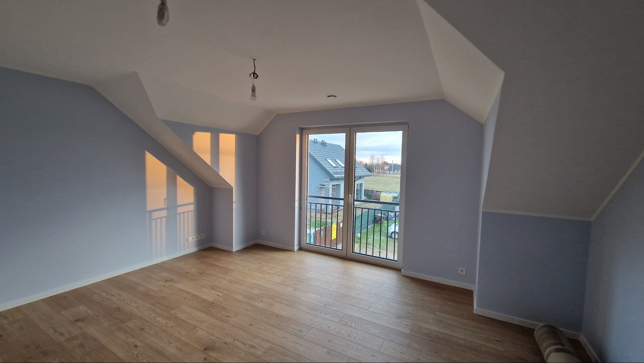 Jasne poddasze z drewnianą podłogą i świeżo pomalowanymi ścianami na delikatny odcień błękitu. Widoczne okno balkonowe z widokiem na sąsiedni dom i ogród. Na suficie widoczne niedokończone...