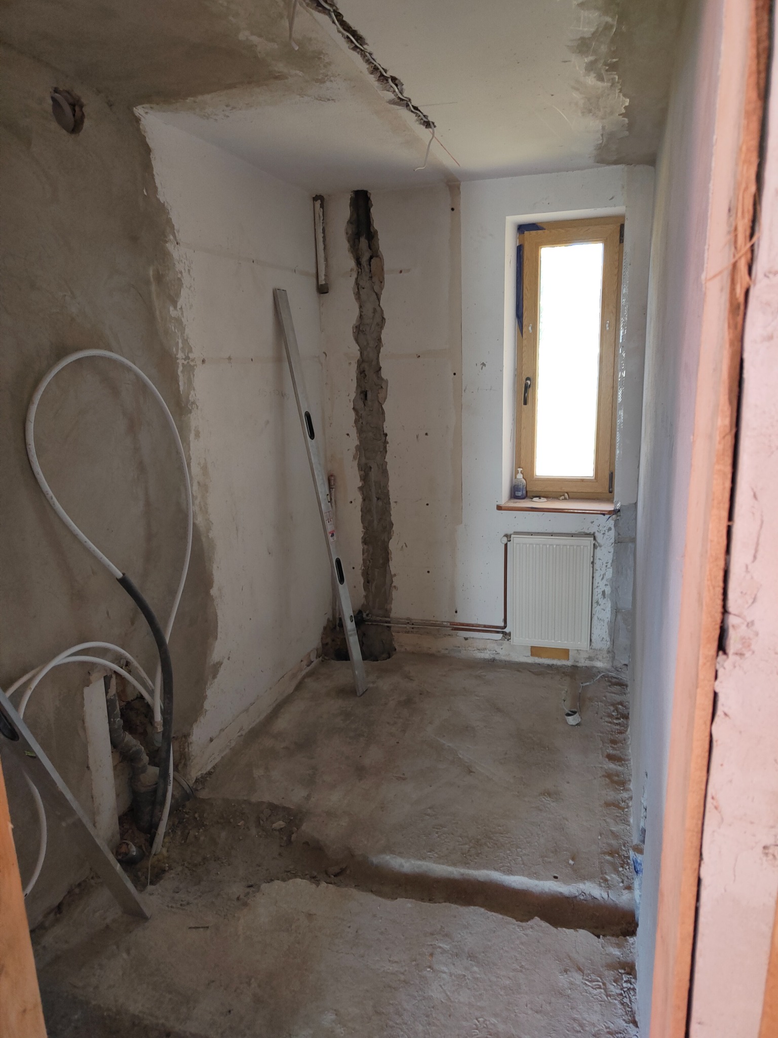 Surowy stan łazienki w trakcie prac: odsłonięte rury, nierówna powierzchnia ścian, poziomica oparta o ścianę, okno z drewnianą ramą i grzejnik pod nim.
