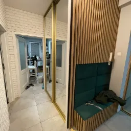 Nowoczesny przedpokój z lustrzaną szafą przesuwną w złotej ramie, ściana z drewnianych listew i tapicerowanym siedziskiem w kolorze butelkowej zieleni, widok na pomieszczenie z białymi meblami.