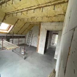 Poddasze w trakcie ocieplania wełną mineralną, widoczne elementy konstrukcji dachu, rusztowanie i ściany z białego bloczka.