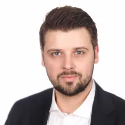 Portret prawnika w czarnym garniturze i białej koszuli na białym tle, z delikatnym uśmiechem i niebieskimi oczami.