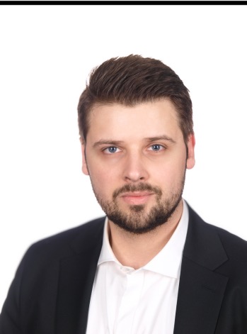 Portret prawnika w czarnym garniturze i białej koszuli na białym tle, z delikatnym uśmiechem i niebieskimi oczami.